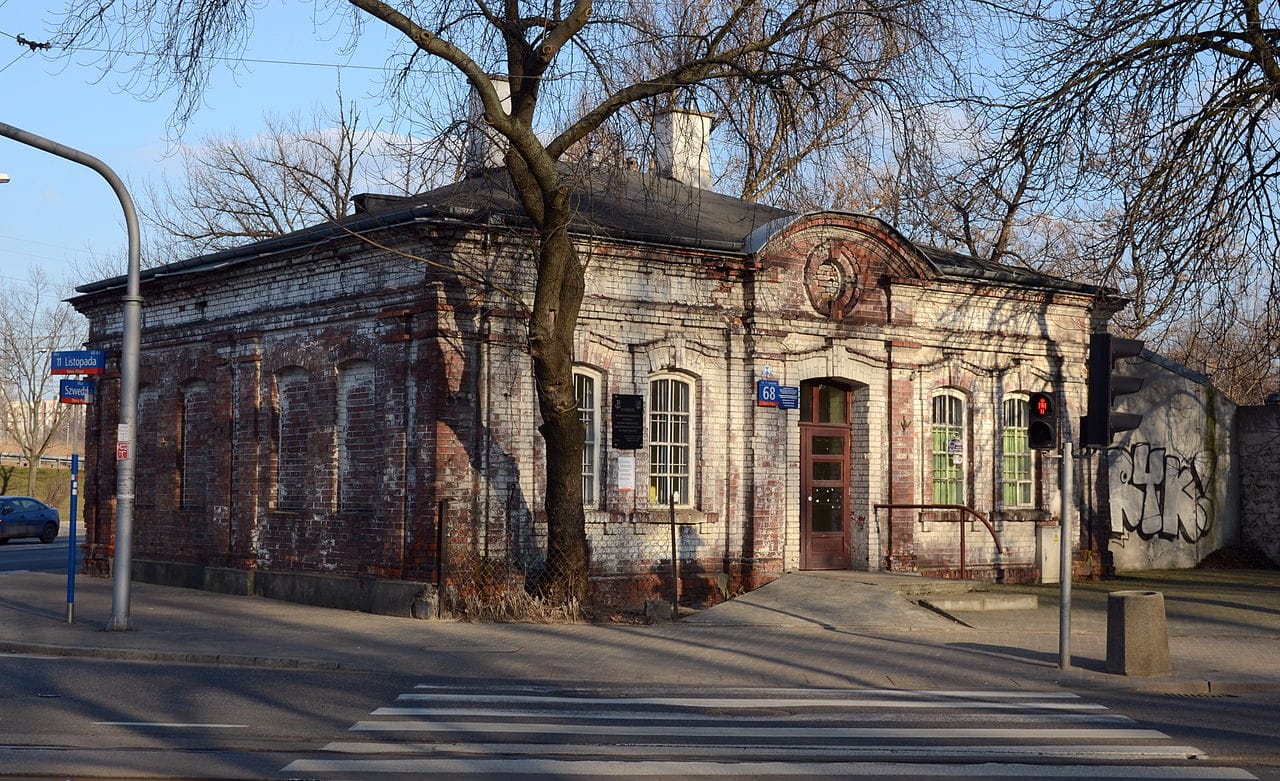 Stary ceglany budynek na rogu ulicy miejskiej, pokazujący ślady wieku i zużycia. Widoczny jest numer 68.