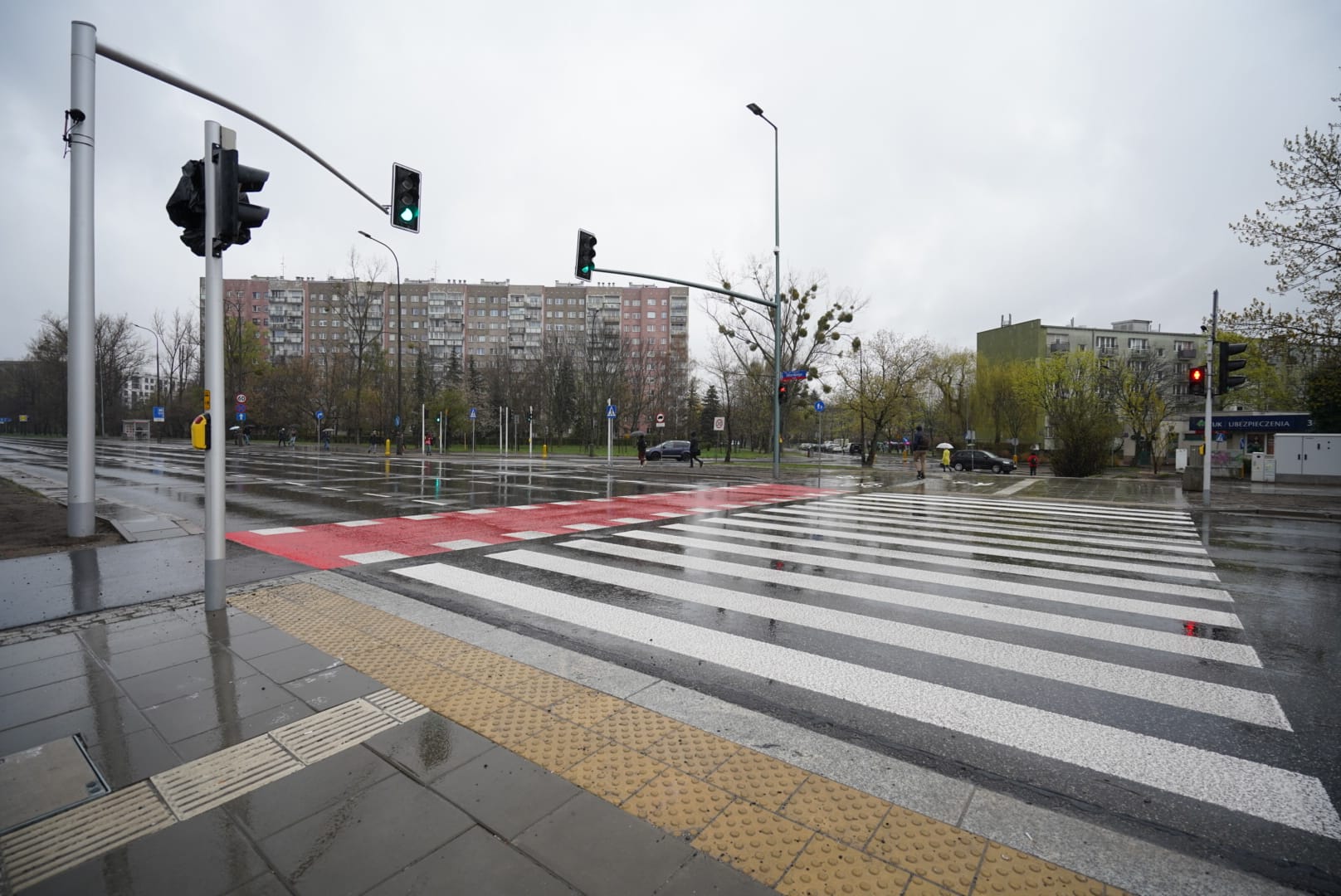 Przejście dla pieszych w deszczowy dzień. W tle widać sygnalizację świetlną i budynki mieszkalne.