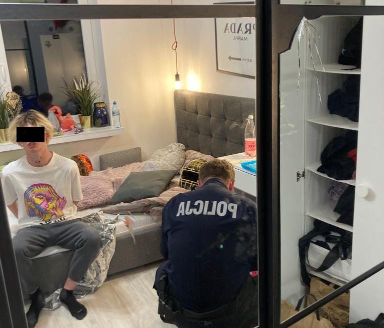 Policjant zatrzymuje młodą osobę w sypialni. Na mundurze policjanta widoczny jest napis POLICJA.