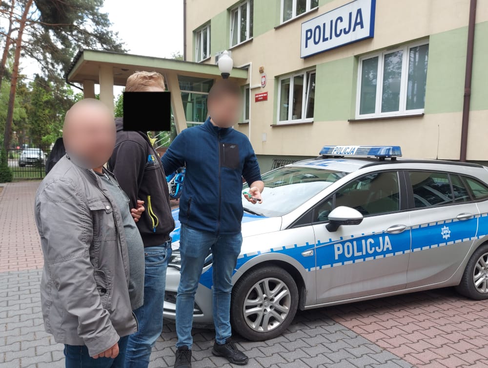 Polscy policjanci zatrzymują dwóch podejrzanych w pobliżu radiowozu. Na radiowozie widać napis POLICJA.