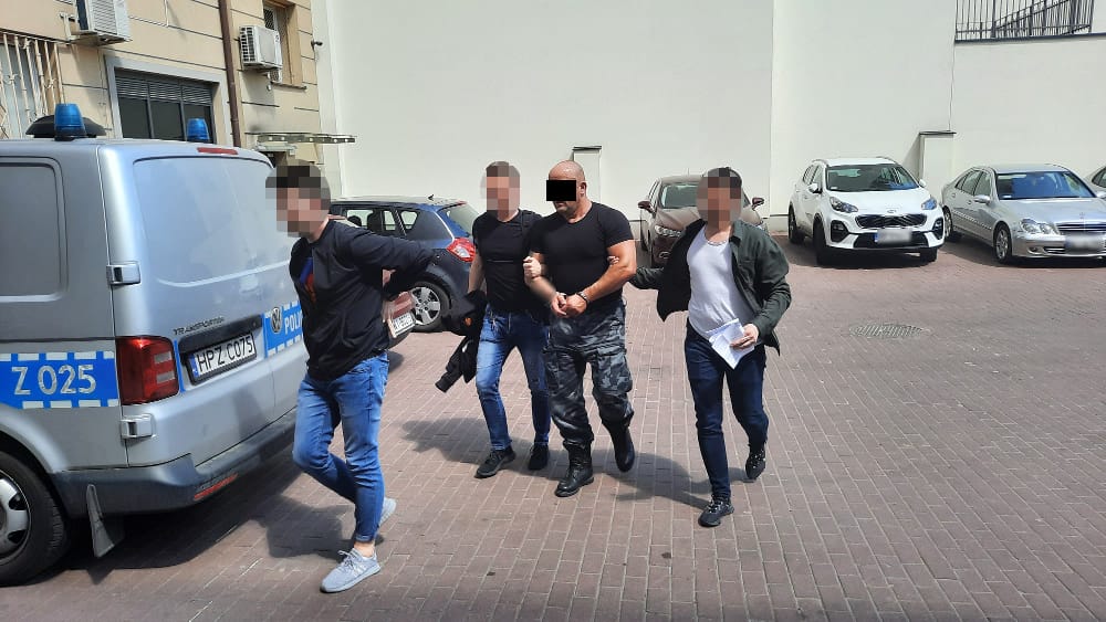 Policjanci eskortują czterech podejrzanych w kajdankach w pobliżu radiowozu.