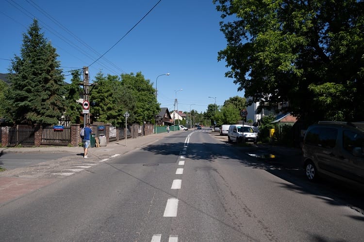 Scena uliczna na terenie osiedli mieszkaniowych w Polsce, ukazująca domy, zaparkowane samochody i pieszego.