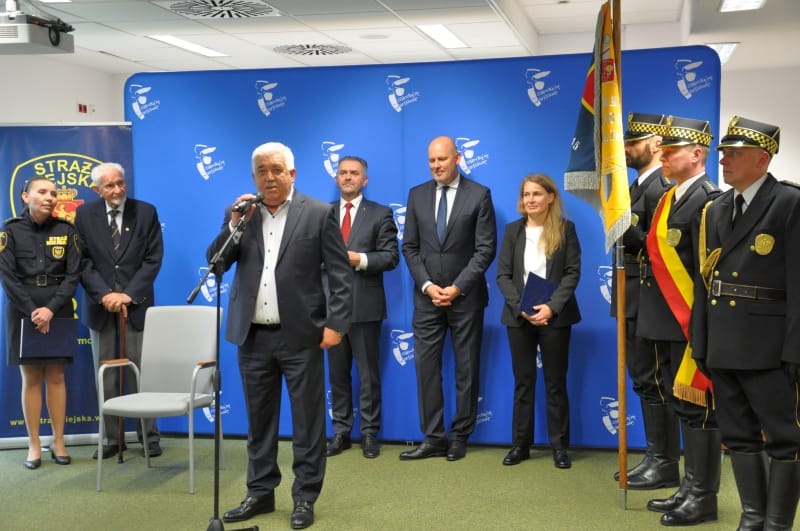 Funkcjonariusze policji miejskiej stoją na baczność podczas ceremonii. Oficjalne władze i goście przyglądają się.