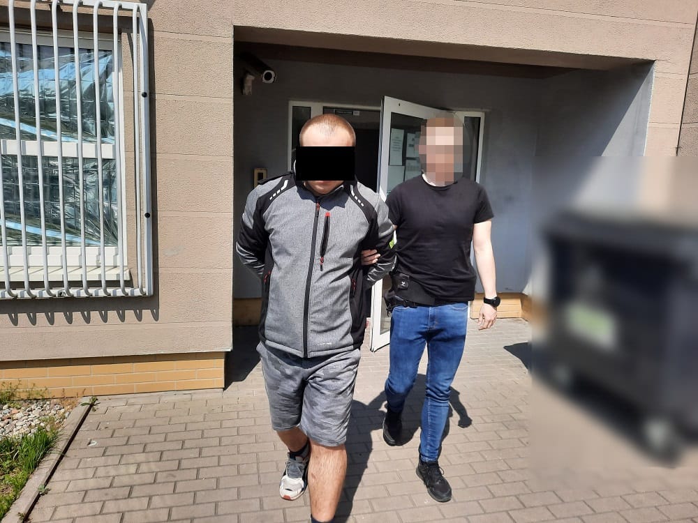 Dwóch mężczyzn w areszcie, eskortowanych przez policję przed budynkiem.