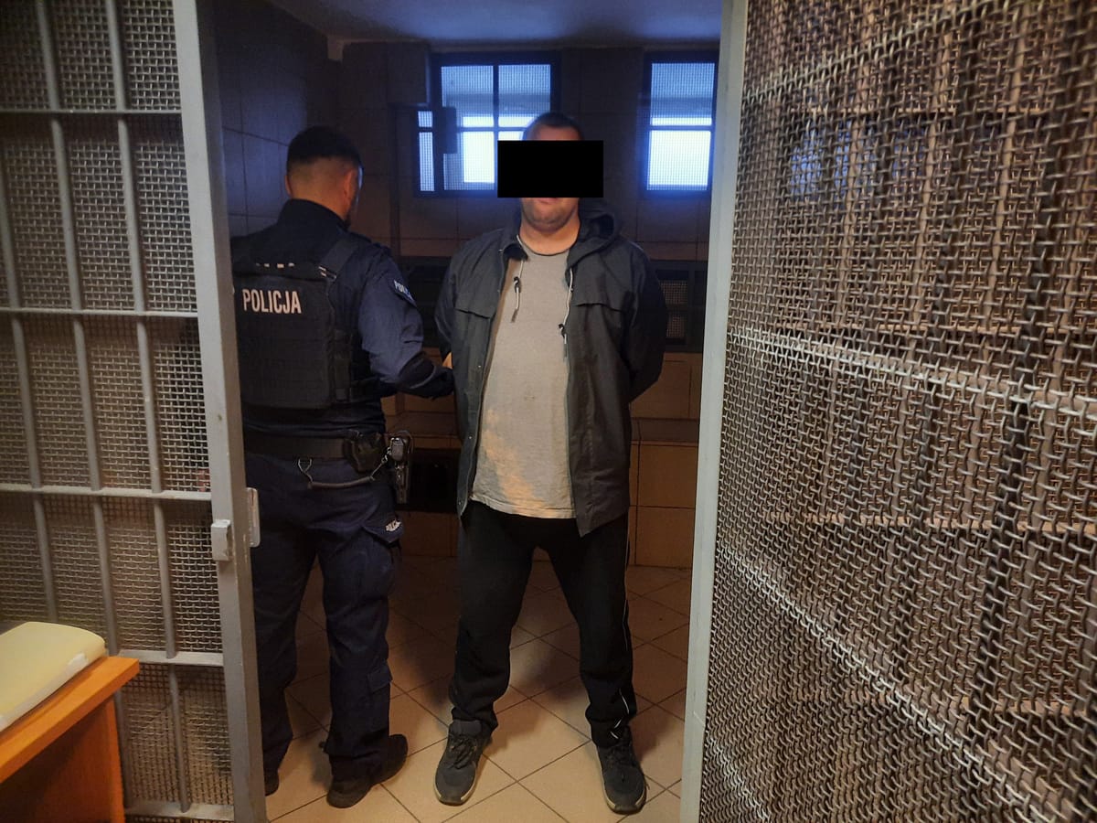 Policjant aresztujący podejrzanego w celi więziennej. Na mundurze policjanta widoczny jest napis „POLICJA”.