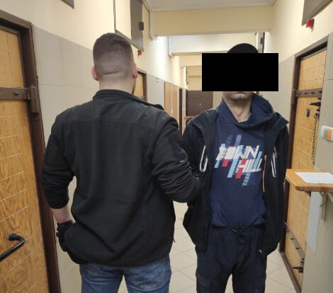 Dwóch mężczyzn w areszcie, jeden z częściowo zasłoniętą twarzą, na korytarzu.