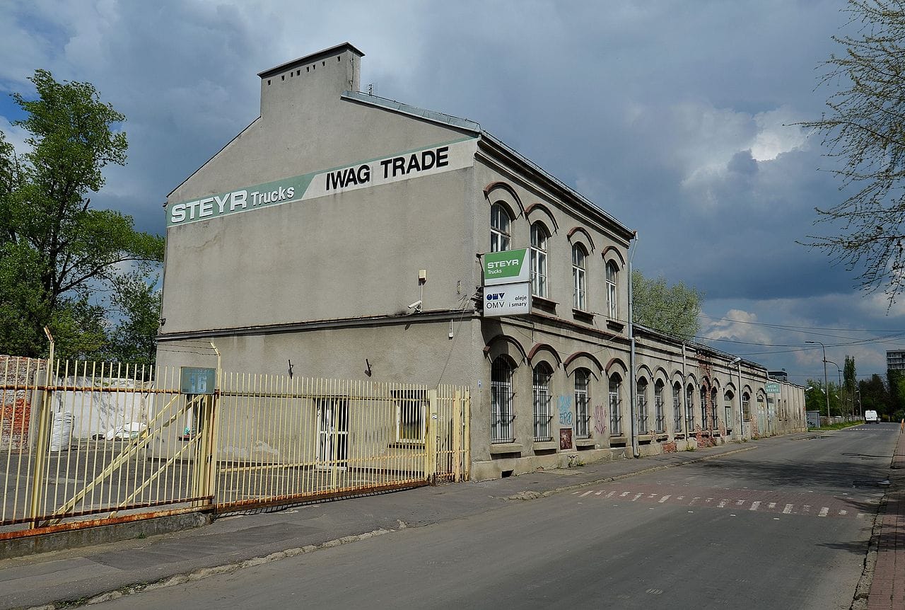 Długi, szary budynek przemysłowy z szyldem „STEYR Trucks” i „IWAG TRADE”.
