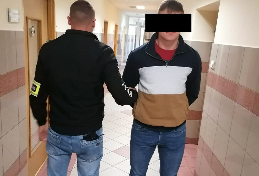 Policjant eskortujący podejrzanego w kajdankach korytarzem.