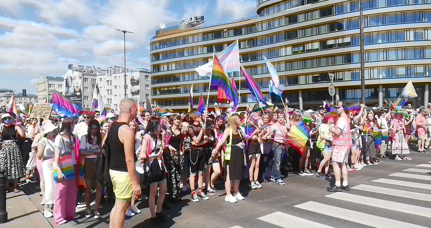 Uczestnicy parady LGBTQ+ maszerują ulicą miasta, trzymając tęczowe flagi i transparenty.