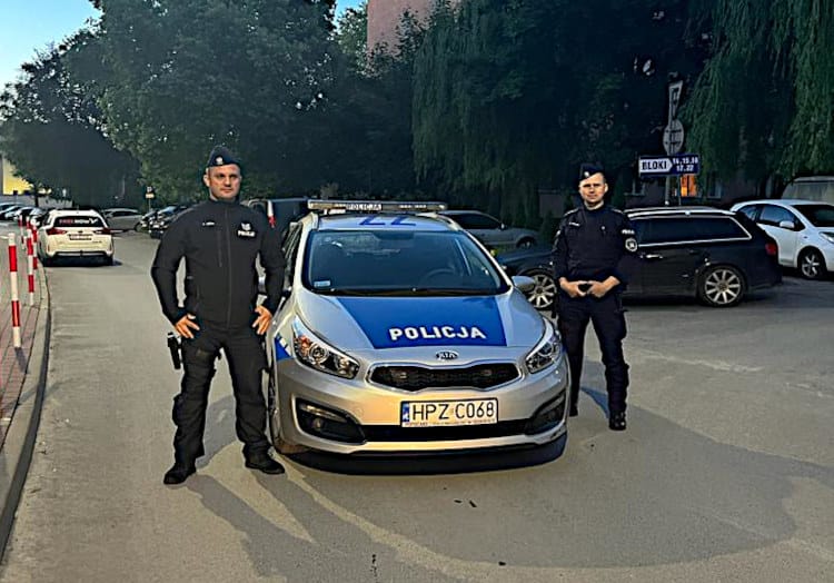 Dwóch polskich policjantów stoi dumnie przy swoim radiowozie z napisem „POLICJA”.