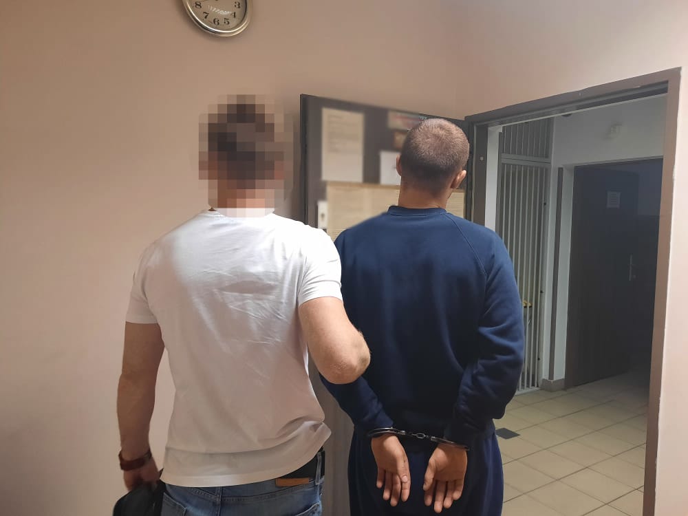 Dwóch mężczyzn w kajdankach, jeden w białej koszuli, drugi w ciemnoniebieskiej bluzie, stoi na korytarzu.