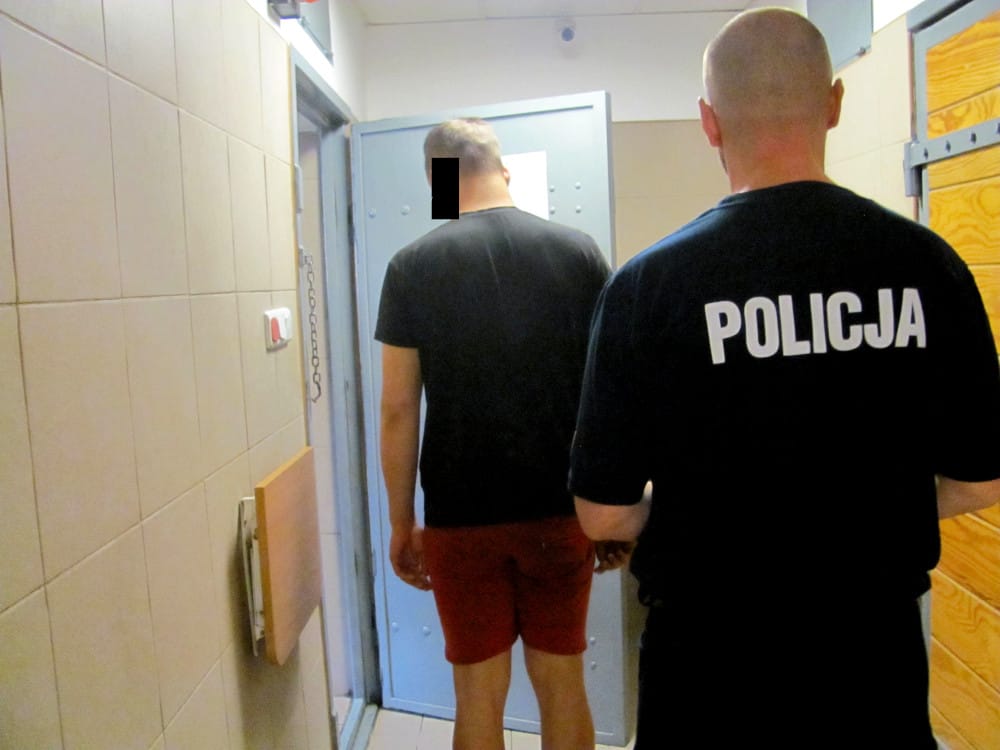 Policjant eskortujący podejrzanego korytarzem. Na mundurze policjanta widoczny jest napis POLICJA.