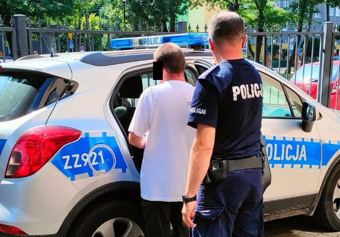 Policjant aresztujący podejrzanego obok radiowozu. Na tablicy rejestracyjnej samochodu widnieje „ZZ921”.