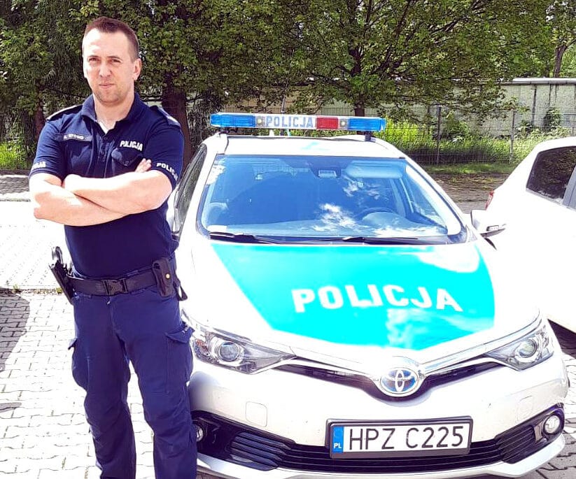 Polski policjant stoi dumnie obok radiowozu z napisem „POLICJA”.