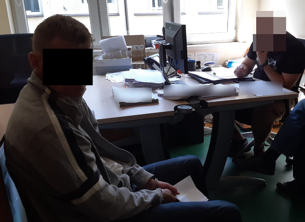 Dwóch podejrzanych w kajdankach siedzi przy stole podczas przesłuchania przez policję. Twarz jednego z podejrzanych jest rozmazana.