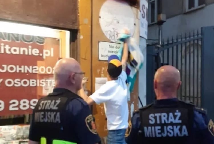 Dwóch strażników miejskich obserwuje mężczyznę zdejmującego plakat ze ściany.