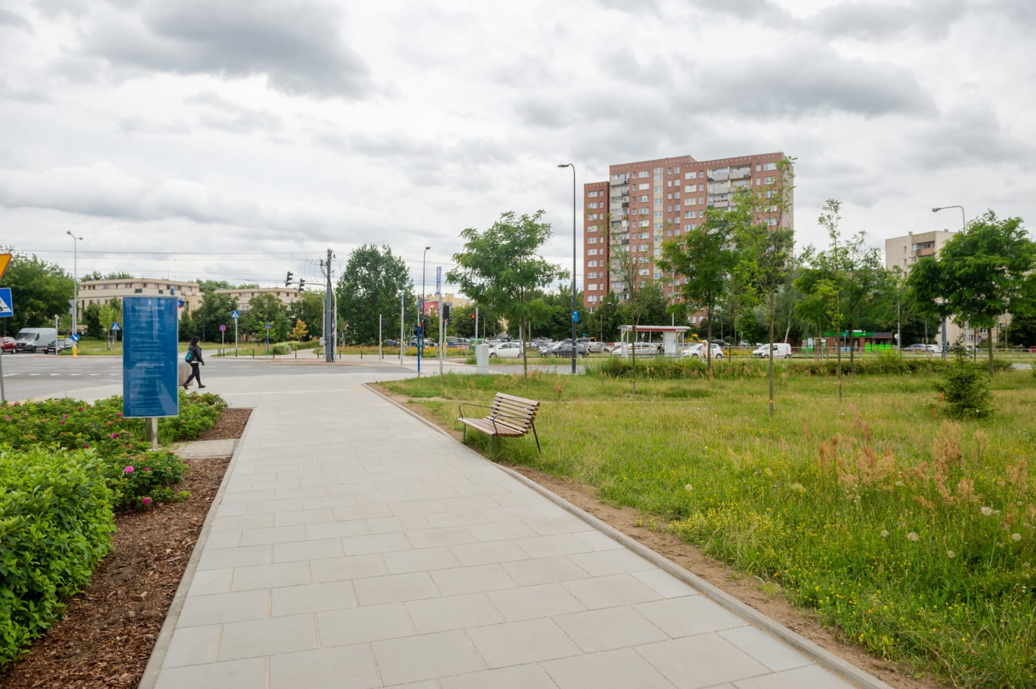 Nowoczesny park miejski z brukowaną alejką, ławką i budynkiem mieszkalnym w tle.