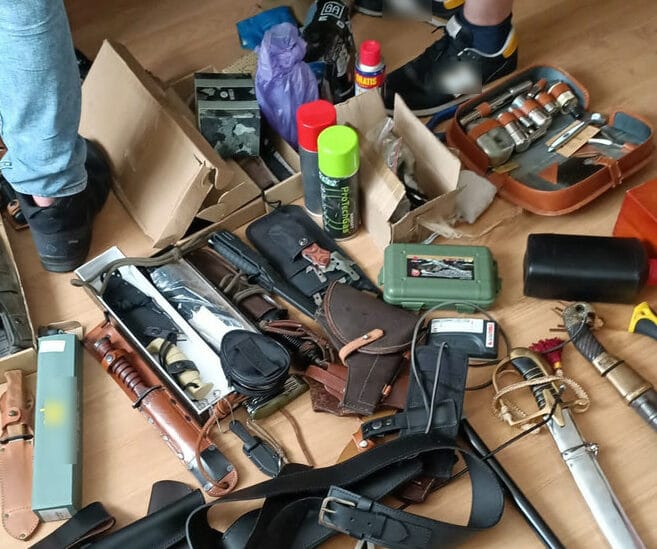 Różne noże, kabury i inny sprzęt taktyczny porozrzucane na drewnianej podłodze.