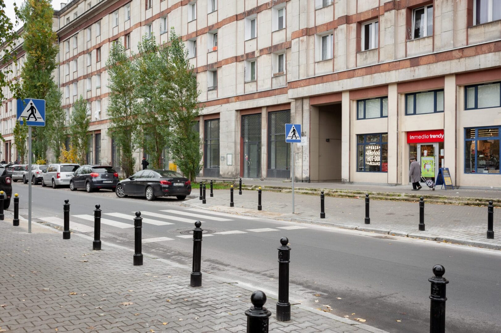 Scena uliczna miasta z zaparkowanymi samochodami, przejściem dla pieszych i sklepem Inmedio Trendy.