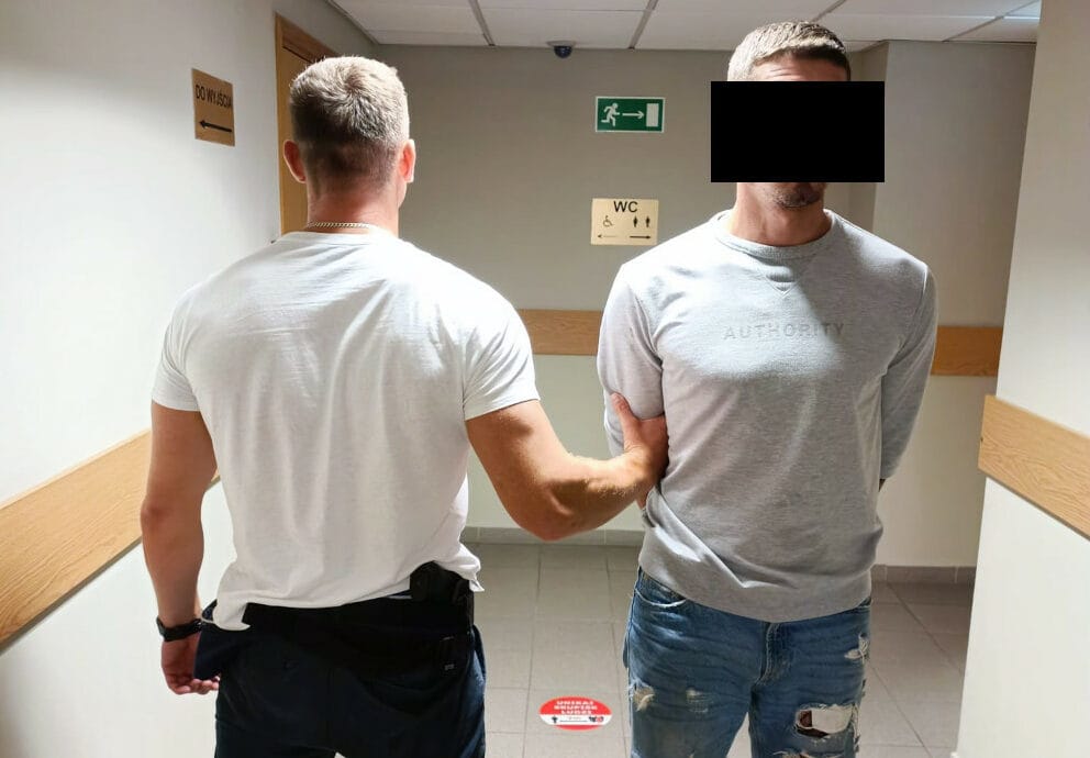 Policjant aresztujący podejrzanego na korytarzu. Na bluzie podejrzanego widoczny jest napis „AUTHORITY”.