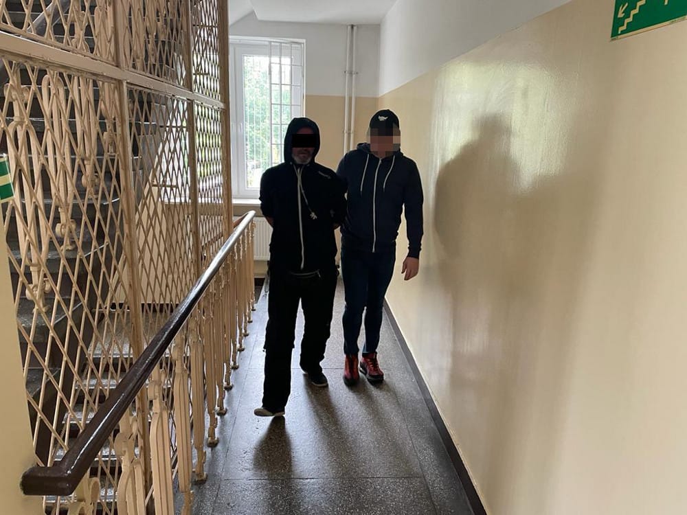 Dwóch mężczyzn w kajdankach idzie korytarzem, eskortowanych przez funkcjonariuszy organów ścigania.