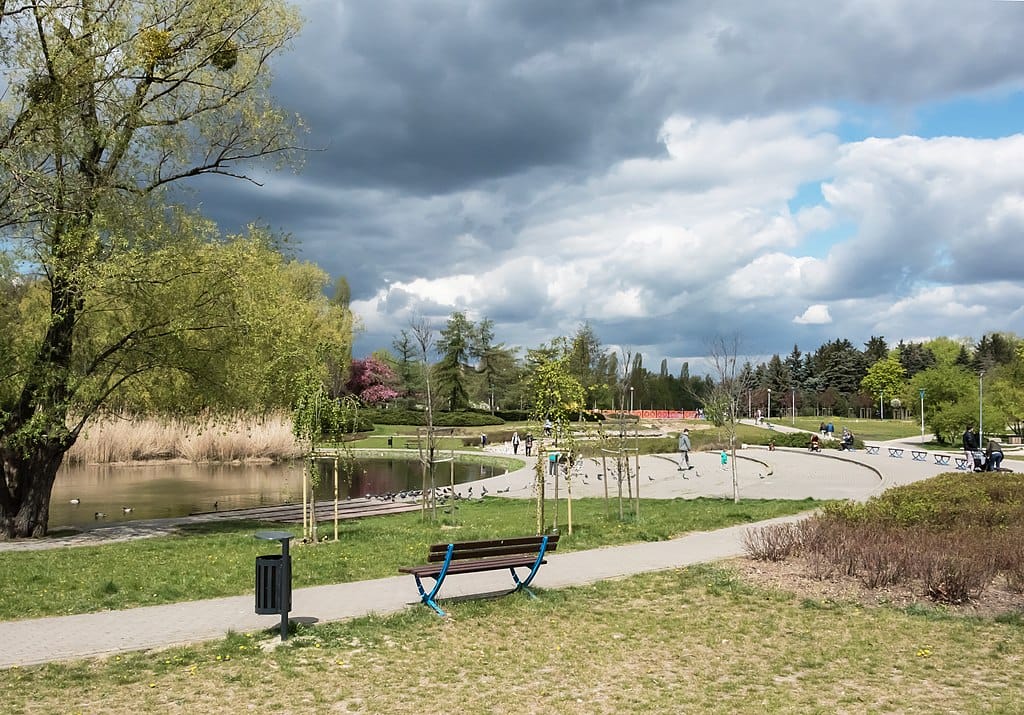 Spokojny park ze stawem, ścieżkami spacerowymi i ławkami pod częściowo zachmurzonym niebem.