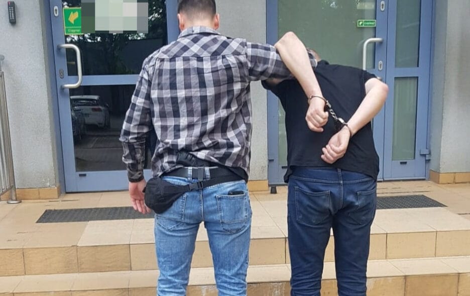 Policjant aresztujący podejrzanego w kajdankach przed budynkiem.