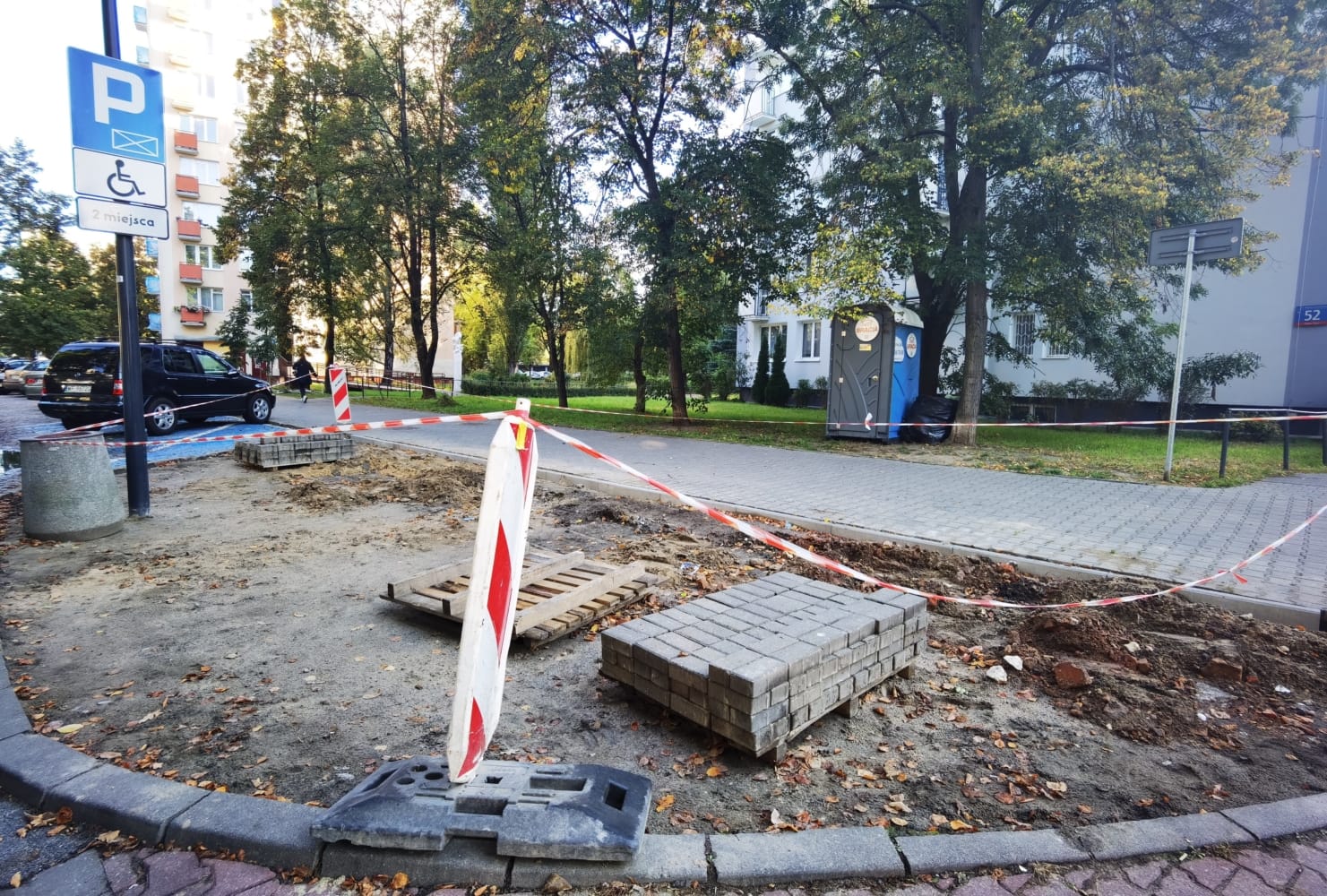 Plac budowy z kostką brukową, prace drogowe i znak informujący o miejscu parkingowym dla niepełnosprawnych.