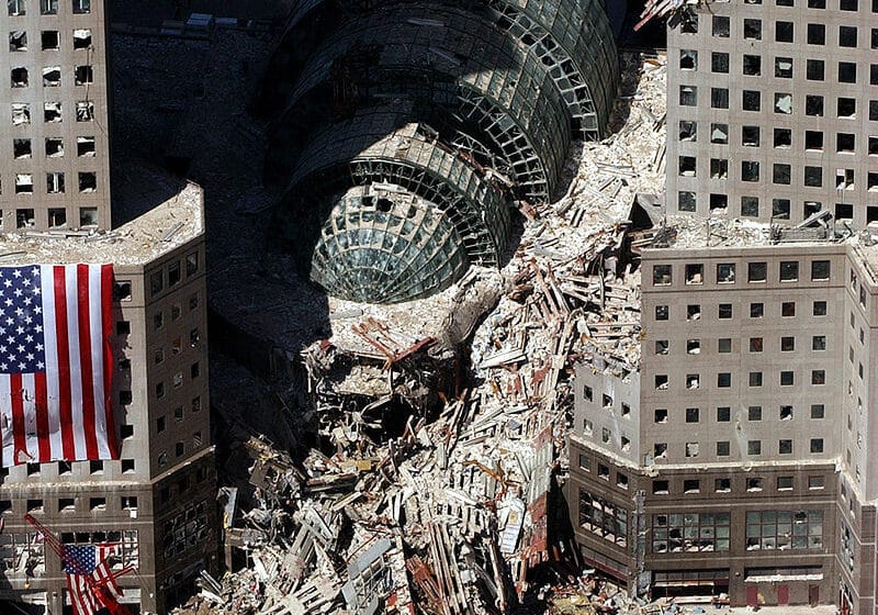 Widok lotniczy gruzów World Trade Center po atakach z 11 września. Widać zniszczone budynki i amerykańską flagę.