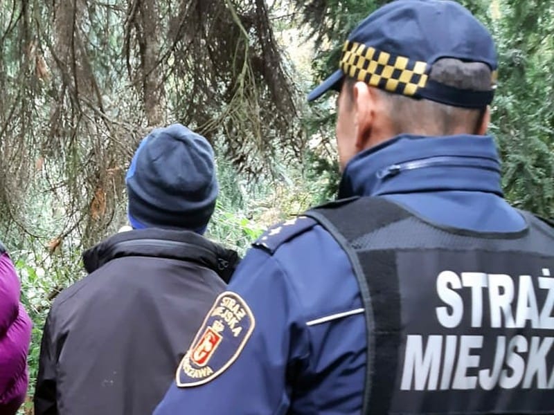 Oficer straży miejskiej i cywile w zalesionym terenie. Na mundurze oficera widoczny napis „Straż Miejska”.