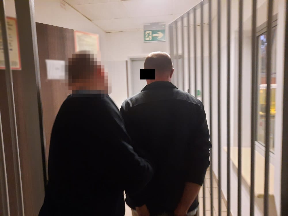 Dwóch mężczyzn w areszcie, idących korytarzem więziennym.