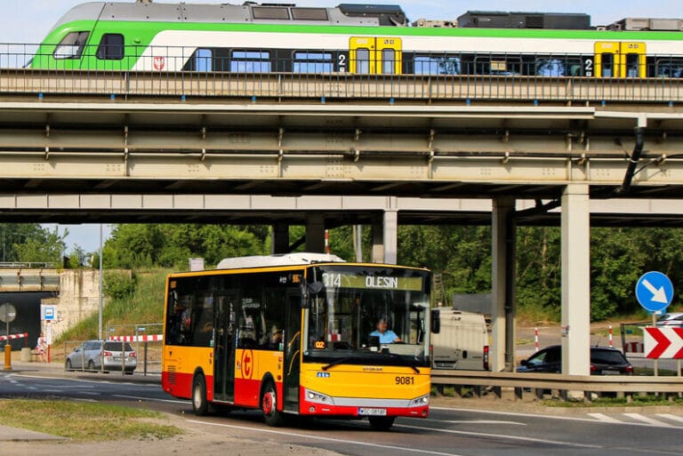 Żółty autobus miejski na drodze pod wiaduktem kolejowym; widoczna stacja docelowa "Olesin".