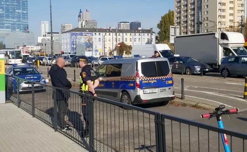 Dwóch policjantów rozmawia przy radiowozie i samochodzie na tle miejskiego krajobrazu.