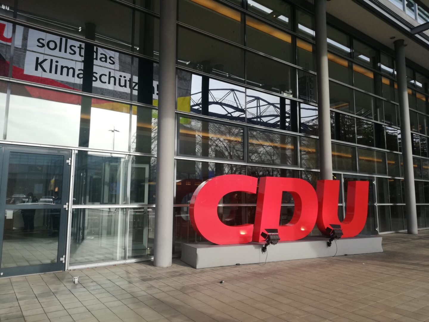 Duże czerwone litery CDU na zewnątrz nowoczesnego budynku. Widoczny baner „sollst das Klimaschutz”.