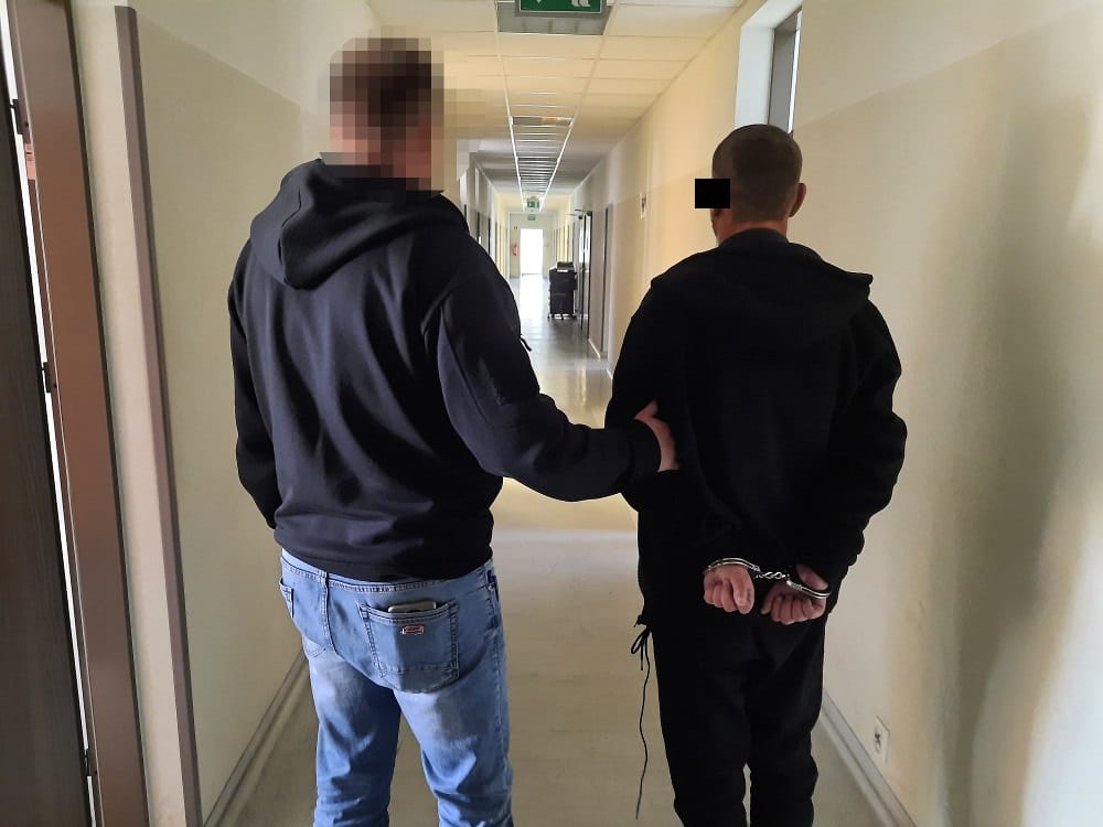 Policjant eskortujący skutego kajdankami podejrzanego korytarzem.