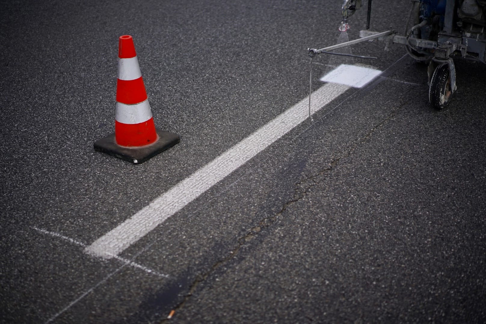 Maszyna do znakowania dróg maluje białą linię na asfalcie, w pobliżu stoi pachołek drogowy.