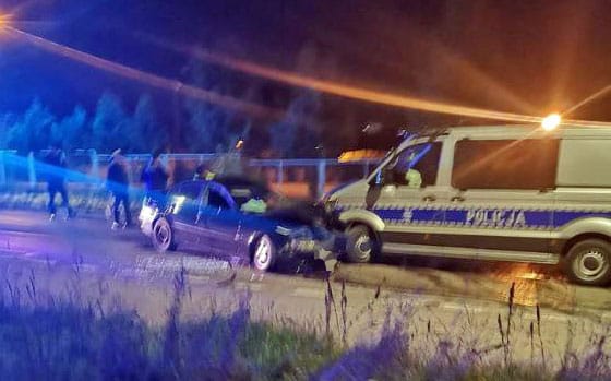 Policja bada wypadek samochodowy w nocy. Widoczny jest uszkodzony samochód i policyjny van.