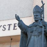 Brązowa statua papieża Jana Pawła II z pastorałem, przed budynkiem, na którym widoczny jest napis „CHRYS”.