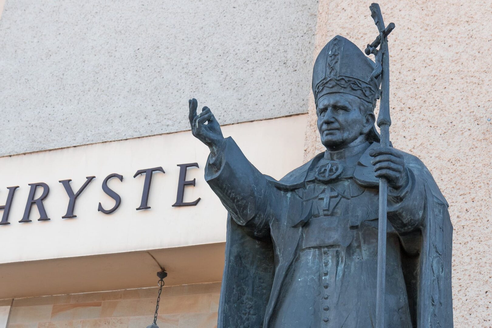 Brązowa statua papieża Jana Pawła II z pastorałem, przed budynkiem, na którym widoczny jest napis „CHRYS”.