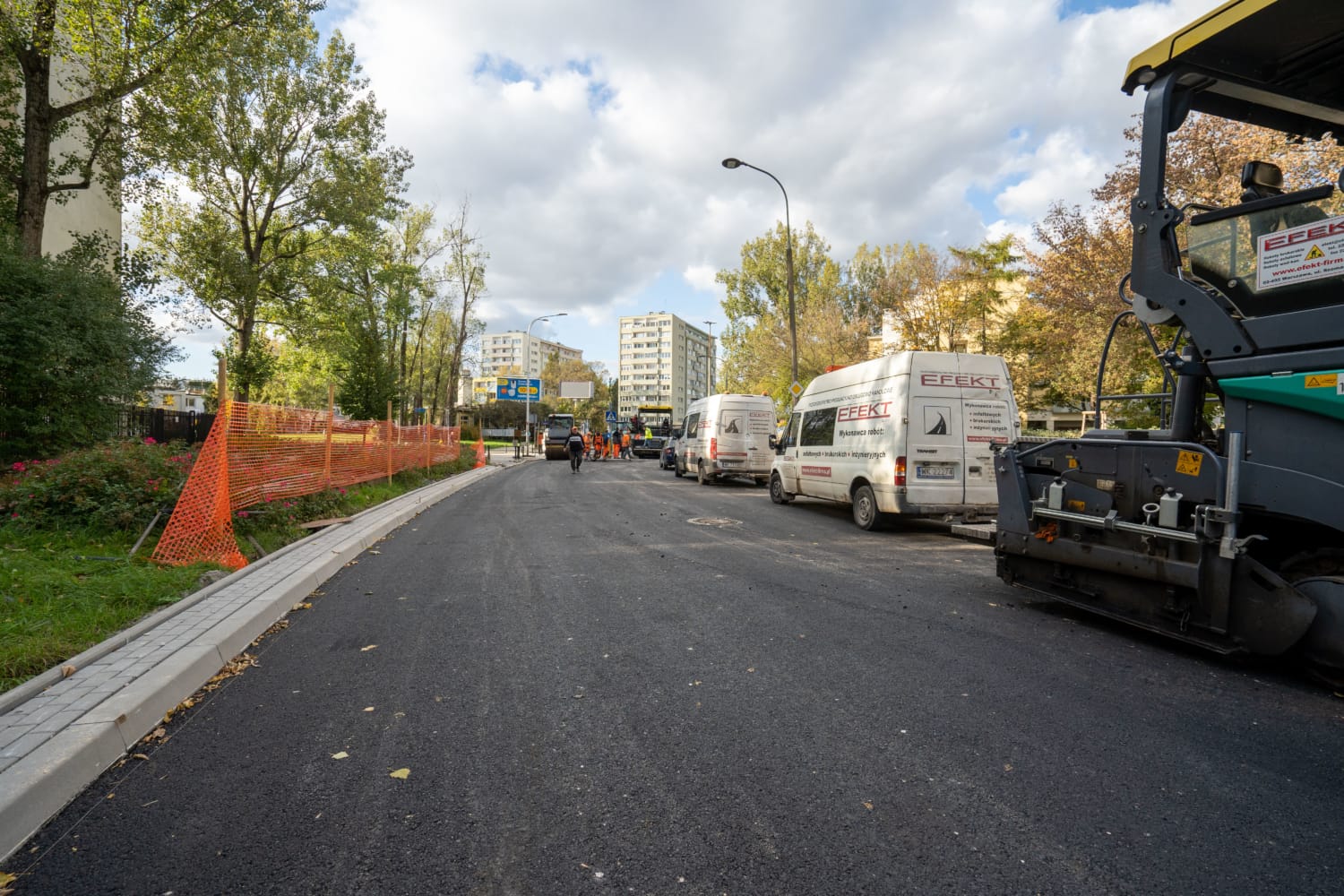 Ekipa drogowa układa nową drogę asfaltową maszyną do układania nawierzchni. W pobliżu zaparkowane są białe vany.