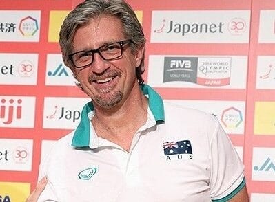 Uśmiechnięty australijski trener siatkówki, ubrany w białą koszulkę polo z australijską flagą.