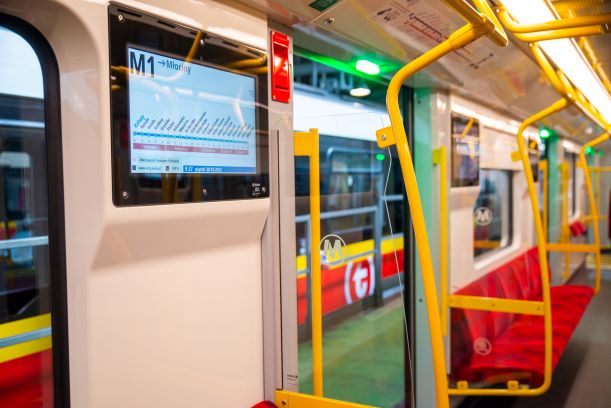 Wnętrze nowoczesnego pociągu metra w Warszawie, linia M1 do Młocin, widoczne siedzenia pasażerów i wyświetlacz cyfrowy.