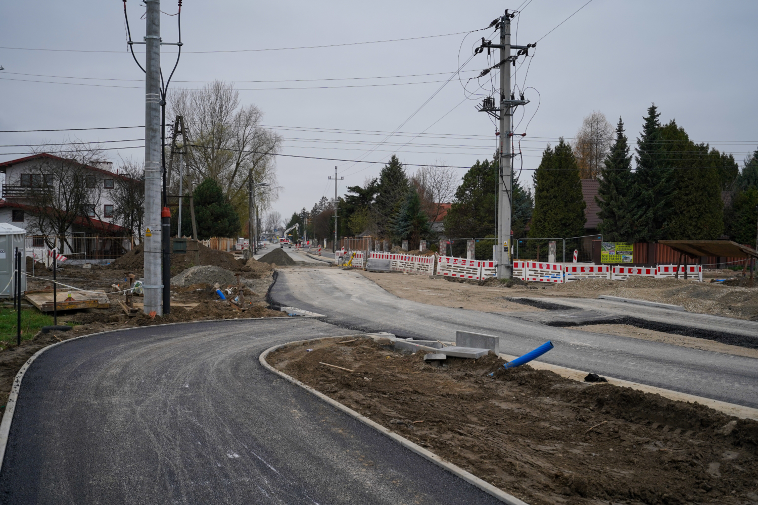Plac budowy drogi z nowym asfaltem, słupami energetycznymi i tymczasowym ogrodzeniem.