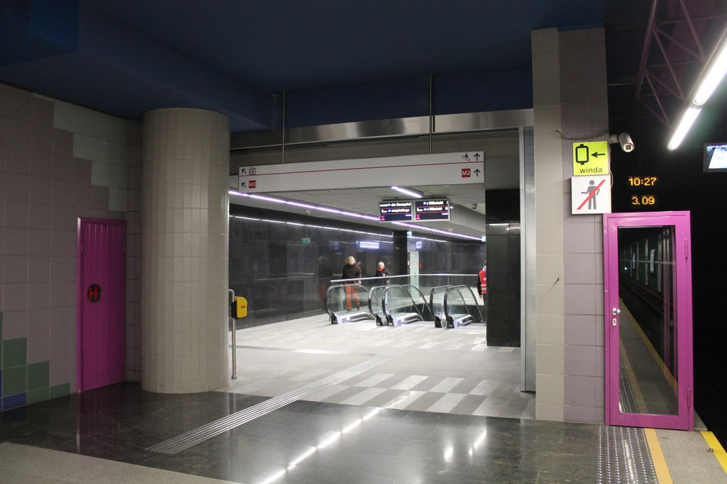 Nowoczesny peron stacji metra z ruchomymi schodami i oznakowaniem wskazującym linię M2.