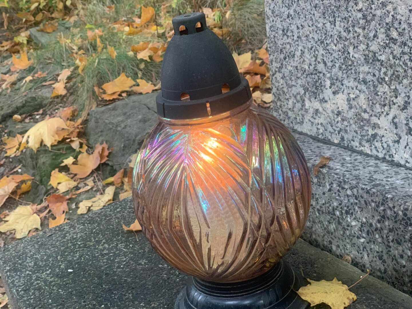 Zapalona szklana świeca wotywna stoi na granitowym nagrobku otoczonym jesiennymi liśćmi.