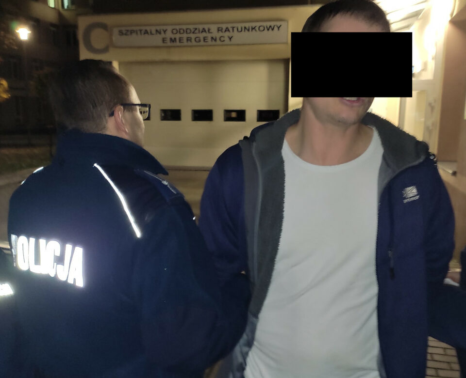 Policjant aresztujący mężczyznę przed szpitalnym oddziałem ratunkowym. Na mundurze policjanta widoczny jest napis „POLICJA”.