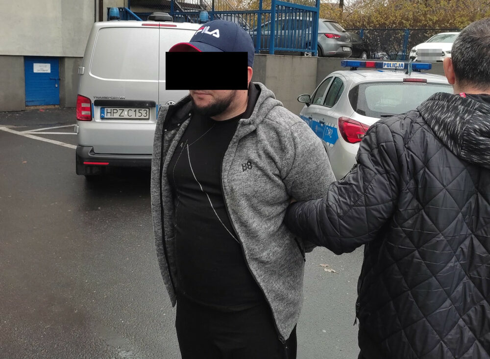 Mężczyzna w kajdankach aresztowany przez policjanta. Widoczna tablica rejestracyjna HPZ C153.