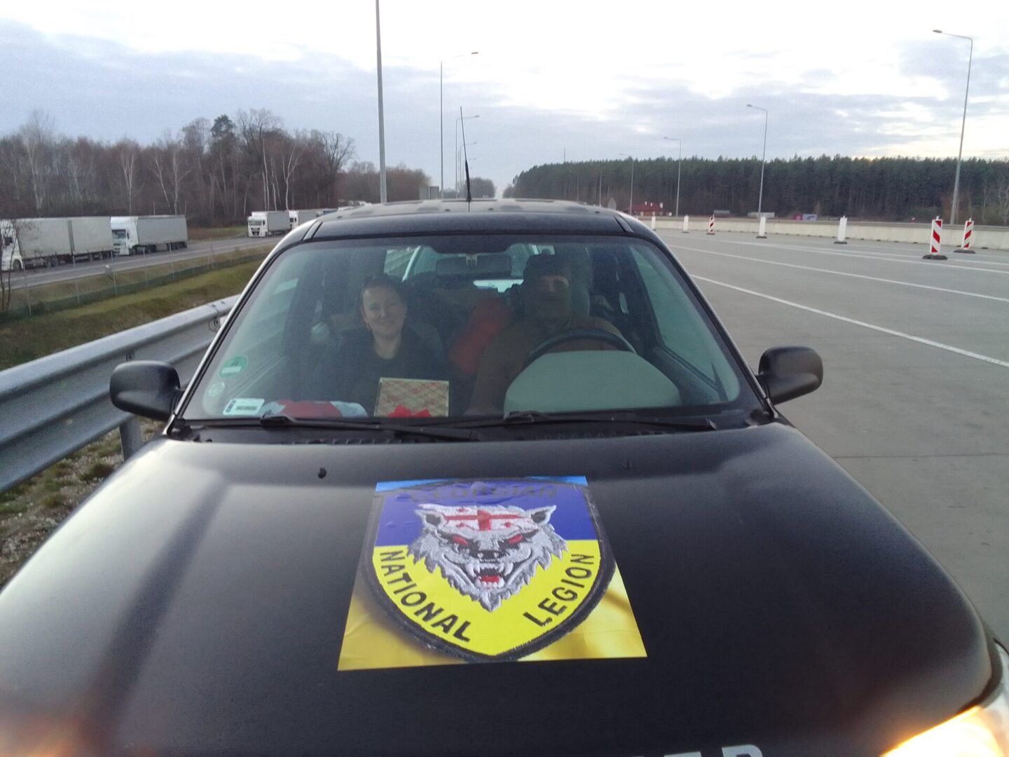 Dwie osoby w samochodzie z emblematem Legionu Narodowego na masce.