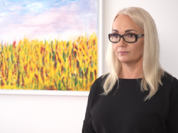 Blondynka w czarnym topie i okularach stoi przed jaskrawym, żółto-zielonym, abstrakcyjnym obrazem.