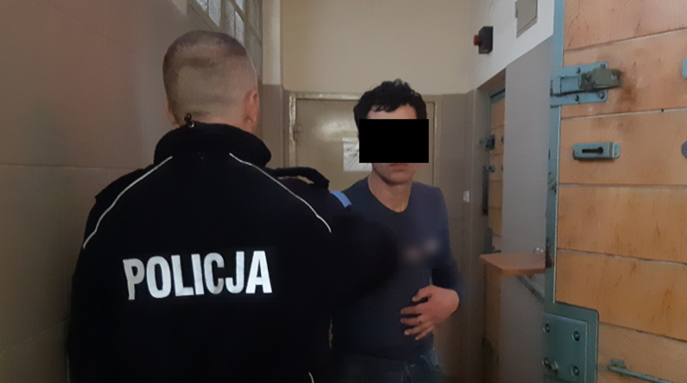 Policjant aresztujący podejrzanego na korytarzu więziennym. Na mundurze policjanta widnieje napis „POLICJA.